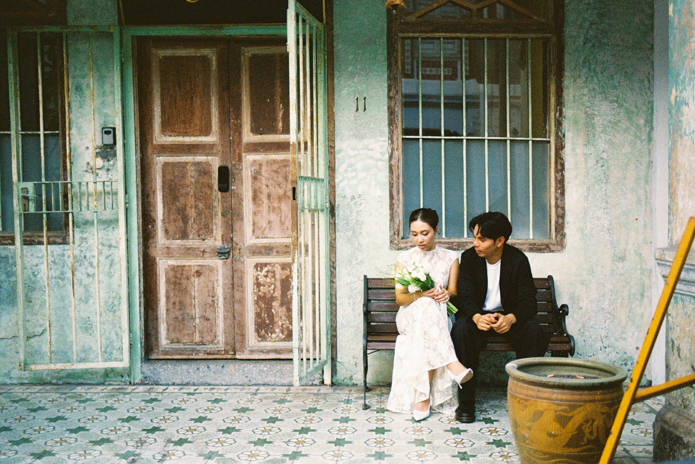 Shafiq and Syahira seated on a bench in front of a weathered teal shophouse with tall wooden doors.