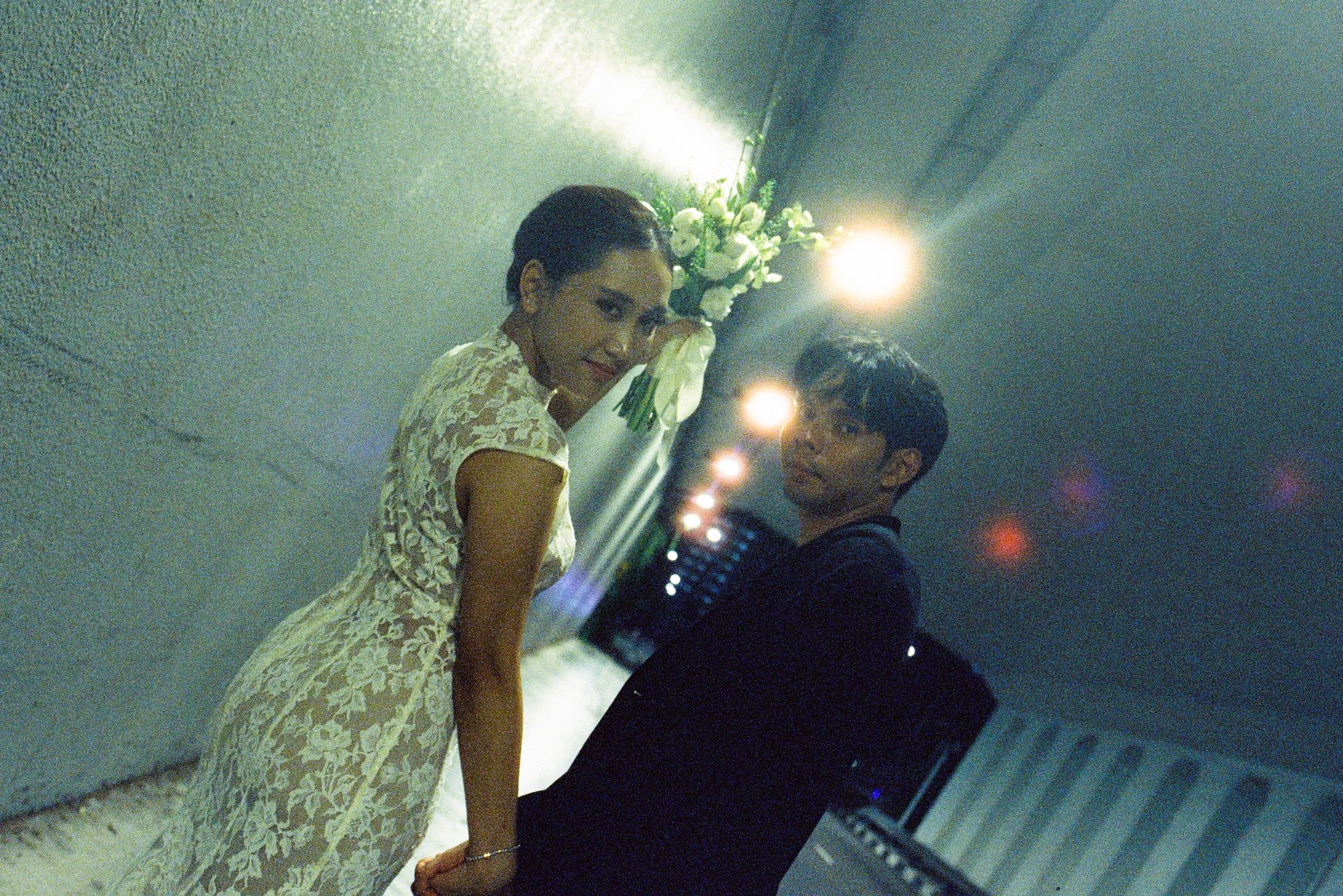 Shafiq and Syahira lit by flash under an overpass at night, Syahira leaning back against the wall with her bouquet raised, lens flares across the frame.